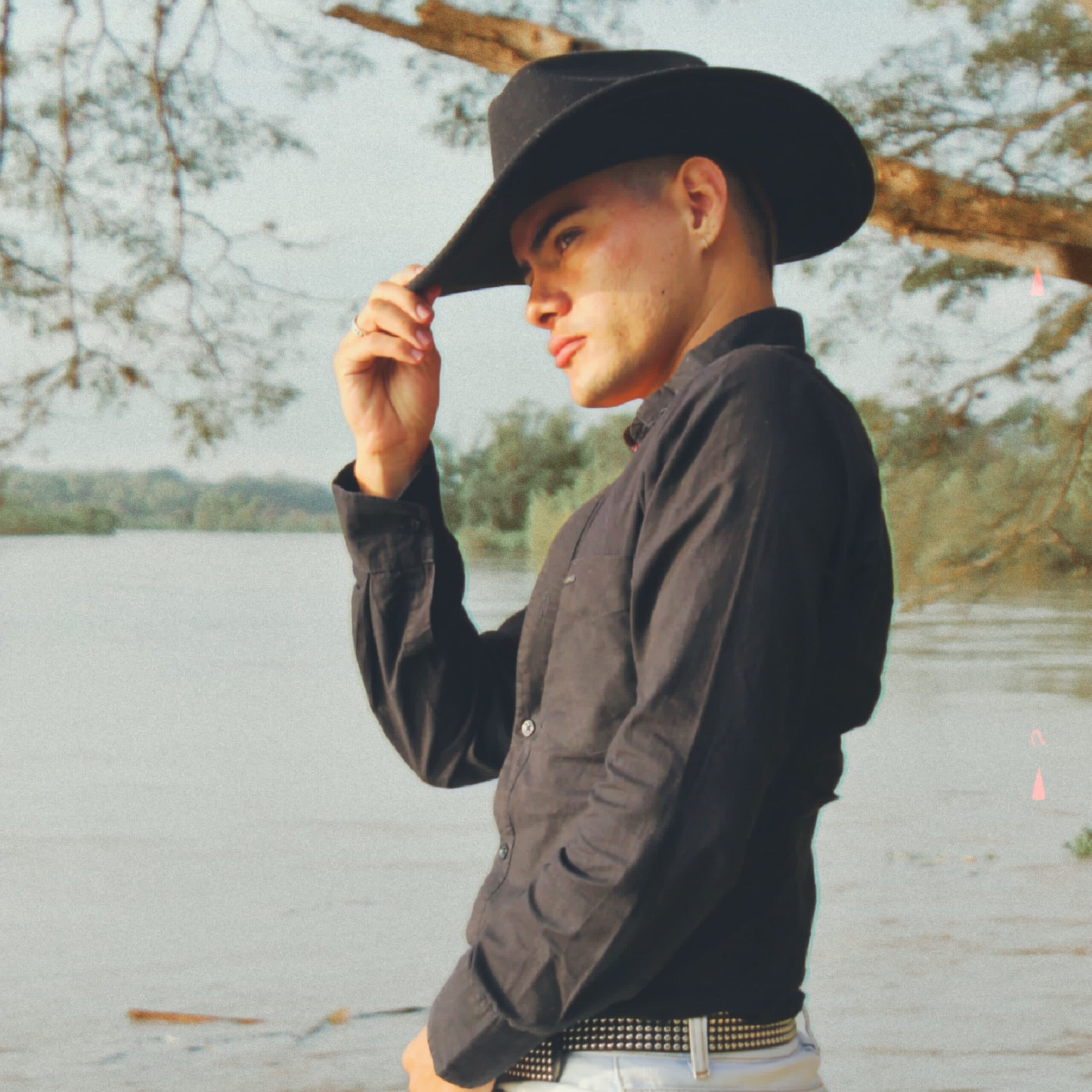 Liam Kai wearing a black cowboy hat, black dress shirt and white jeans standing in front of a wide river